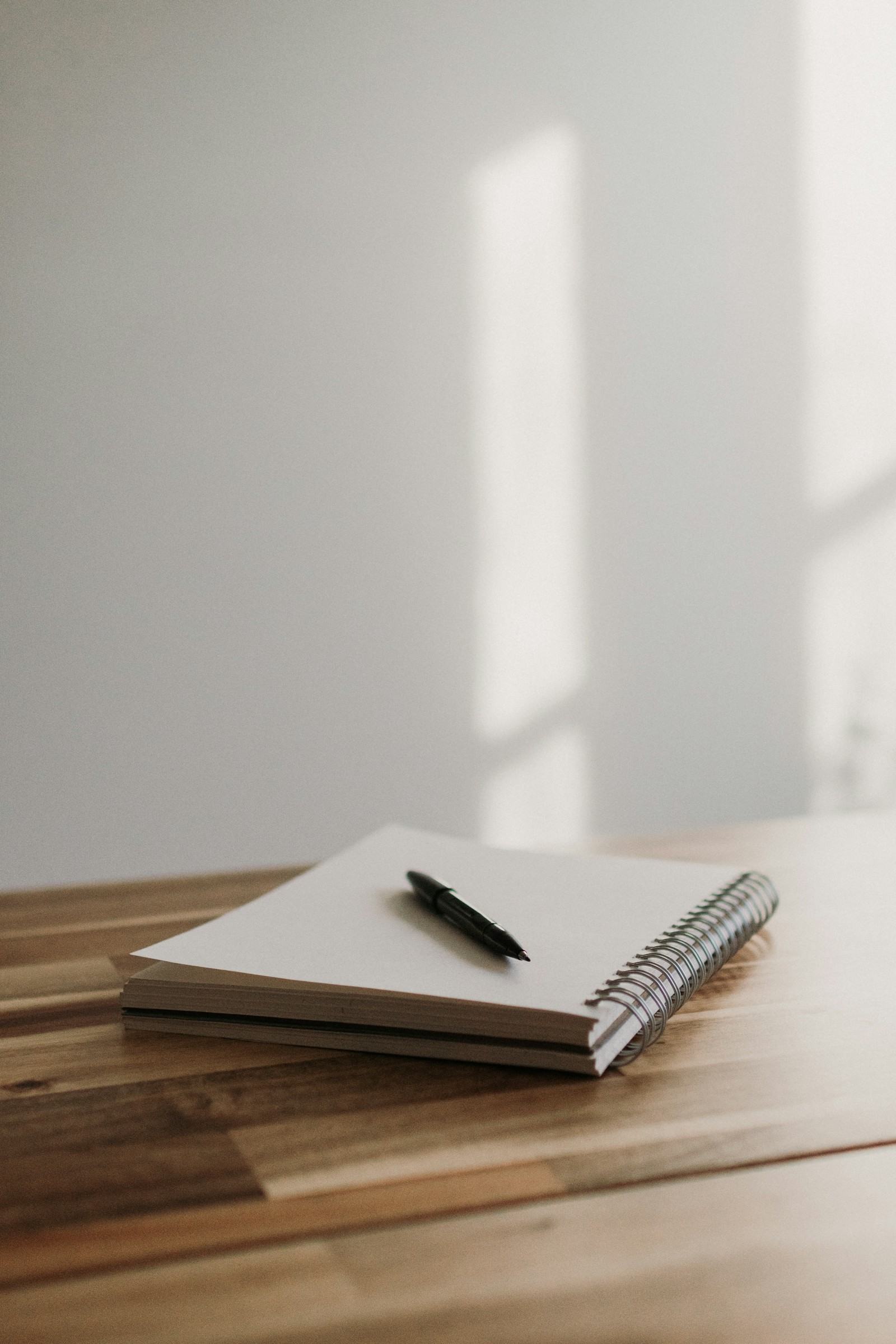 A closed notebook and pen on a wooden table in soft window light